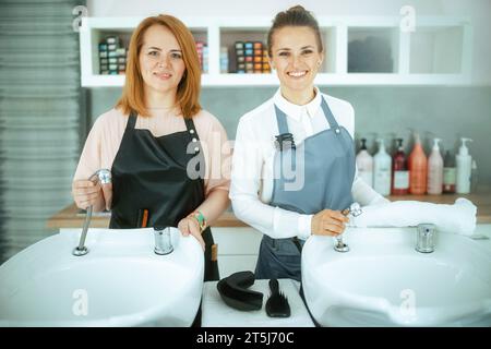 Portrait de souriant deux femmes d'âge moyen employés de salon de coiffure dans des tabliers dans le studio de coiffure moderne près de l'unité de lavage à contre-courant de salon. Banque D'Images