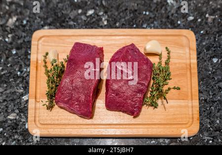 Deux steaks de venaison en préparation pour la cuisson sur planche à découper avec thym et ail Banque D'Images