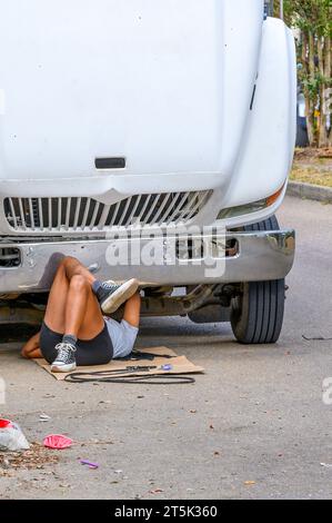 NOUVELLE-ORLÉANS, LA, USA - 29 OCTOBRE 2023 : Femme couchée sur son dos sous l'avant d'un gros camion remplaçant la courroie du ventilateur. Banque D'Images