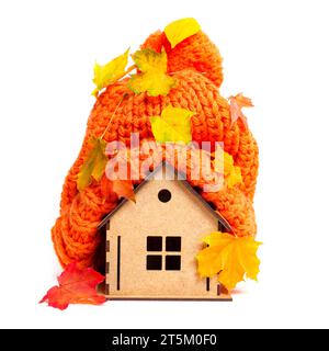 Bonnet en tricot orange chunky recouvert de feuilles pigmentées est placé sur le dessus d'une maison en bois jouet isolé sur blanc. Concept lié à l'isolation de la maison. Banque D'Images