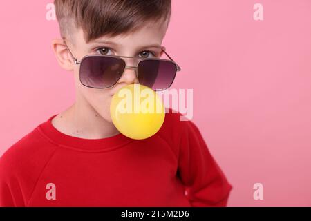 Garçon dans les lunettes de soleil soufflant de la gomme à bulles sur fond rose, espace pour le texte Banque D'Images