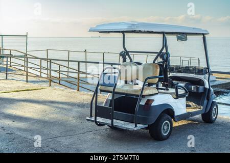 Voiturette de golf confortable garée sur le front de mer de la ville par mer tranquille. Petite voiture touristique sur le remblai en pierre près de la baie de l'océan. Véhicule compact au complexe Banque D'Images