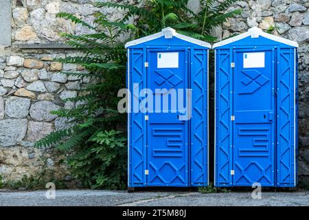 Cabines de WC bio portables publiques par mur de pierre sur la rue de la ville. Toilettes mobiles installées pour les invités dans le parc urbain. Unités sanitaires bleues en ville Banque D'Images