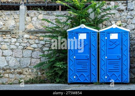 Cabines de WC bio portables publiques par mur de pierre sur la rue de la ville. Toilettes mobiles installées pour les invités dans le parc urbain. Unités sanitaires bleues en ville Banque D'Images