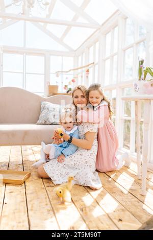 Portrait vertical de mignon jeune maman et deux adorables filles en robe jouant avec de petits canetons jaunes dans la maison gazebo d'été sur la journée ensoleillée. Banque D'Images