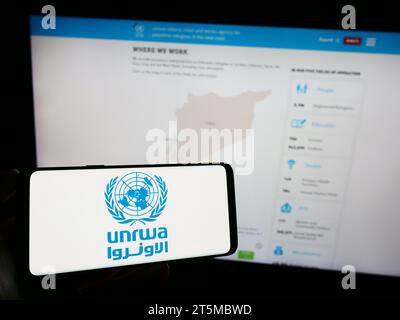 Personne tenant un téléphone portable avec le logo de l'agence des Nations Unies pour les réfugiés Plalestine UNRWA devant la page Web. Concentrez-vous sur l'affichage du téléphone. Banque D'Images