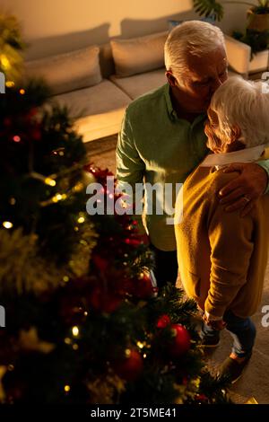 Heureux couple aîné caucasien debout près de l'arbre de noël et embrassant dans le salon à la maison Banque D'Images