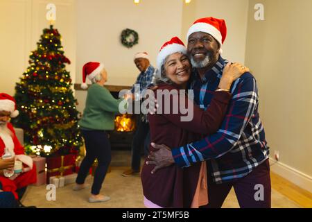 Deux heureux couple senior diversifié d'amis dans les chapeaux de père noël, embrassant dans le salon à la maison. Retraite, noël, célébration, amitié, ensemble Banque D'Images