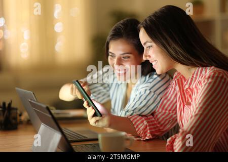 Deux femmes heureuses utilisant plusieurs appareils vérifiant le téléphone intelligent dans la nuit à la maison Banque D'Images