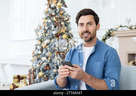 Portrait d'un jeune homme assis à la maison sur le canapé près du sapin de Noël et utilisant un téléphone portable. Regarder et sourire à la caméra. Banque D'Images