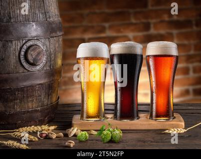 Trois verres réfrigérés de différentes bières et tonneaux de bière sur une table en bois. Banque D'Images