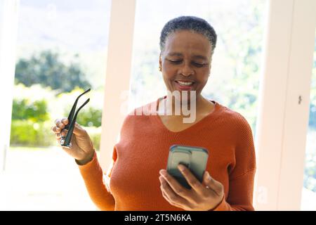 Heureuse femme aînée afro-américaine avec des lunettes, ayant un appel vidéo dans le salon ensoleillé, espace de copie Banque D'Images