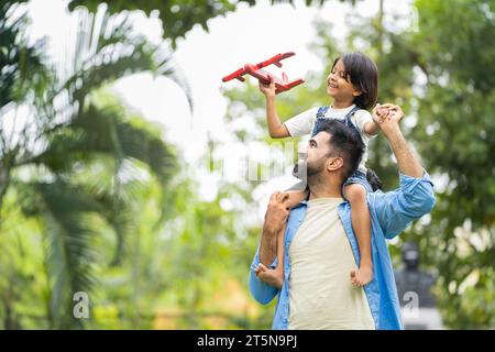 Heureux souriant fille indienne jouant avec le jouet d'avion en s'asseyant sur l'épaule du père au parc - concept de liberté, soutien familial et ensemble Banque D'Images