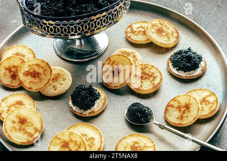 Blinis au caviar noir et fromage à la crème, sur un plat festif sur fond noir, mini crêpes, un élégant apéritif Banque D'Images