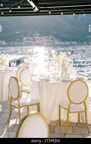 Des chaises blanches se dressent autour de tables de fête rondes avec des bouquets de fleurs sous une couverture sur la jetée Banque D'Images