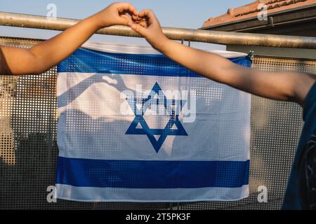 Les mains d'une fille et d'un garçon forment une forme de coeur sur le drapeau israélien, avec la lumière du soleil coulant une ombre sur le drapeau national suspendu au balcon. Banque D'Images