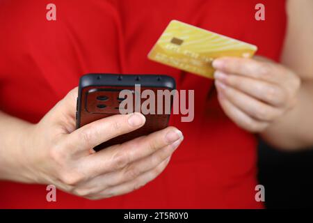 Femme en robe rouge tenant le smartphone et la carte de crédit entre les mains. Concept de paiements, d'achats et de transactions en ligne Banque D'Images