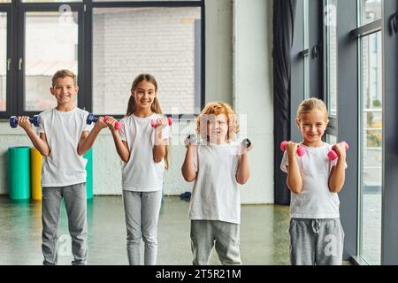 joyeux enfants préadolescents s'exerçant avec des haltères et souriant joyeusement à la caméra, au sport Banque D'Images