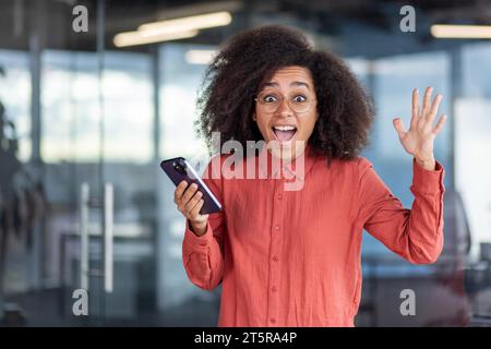 Portrait Joyeuse femme d'affaires gagnante, employé entrepreneur souriant et regardant la caméra, célébrant les réalisations de résultats réussis, tenant le téléphone dans les mains et levant la main dans le geste de triomphe Banque D'Images
