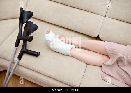 Jeune femme avec fracture de l'os du pied reposant sur le canapé à la maison. Jambe cassée d'adulte dans un plâtre. Béquilles à proximité tout en étant assis sur le canapé Banque D'Images