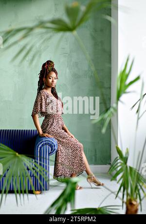 Сonfident jeune femme africaine, vêtue d'une robe avec imprimé léopard, assise sur le bras du canapé et regardant la caméra contre un mur vert menthe. I. Banque D'Images