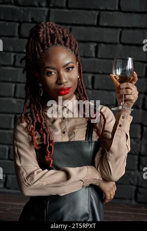 Portrait en gros plan en demi-corps d'une femme africaine confiante et bien habillée tenant un verre de vin blanc sur un fond de brique noire. Dégustation de vins, Banque D'Images