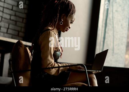 Femme africaine travaillant à l'ordinateur portable. Employé travaillant à distance à la maison tout en étant confortablement assis dans une chaise contre la fenêtre. Bureau de marketing numérique, W Banque D'Images