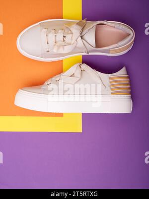 Baskets contemporaines pour femmes beiges décontractées sur un fond violet-jaune-orange avec espace de copie sur le bas. Chaussures de sport décontractées tendance et stylées Banque D'Images