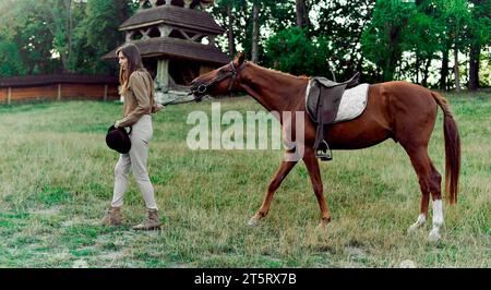 Jeune jockey-fille menant le cheval brun sellé par les rênes sur un terrain d'herbe. École équestre, centre équestre, cours d'équitation. Equitrian prenant ho Banque D'Images