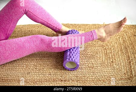 Technique de roulis pour les zones cibles. Jambes féminines fines pieds nus en sportswear rose sur un tapis écologique tissé. Masseur à rouleau en mousse violette étirant les jambes pour se détendre Banque D'Images