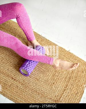 Les jambes des femmes en vêtements de sport roses s'allongent pieds nus sur un tapis écologique tissé. Masseur à rouleau en mousse violette pour détendre et renforcer la force des fessiers. Target sont Banque D'Images