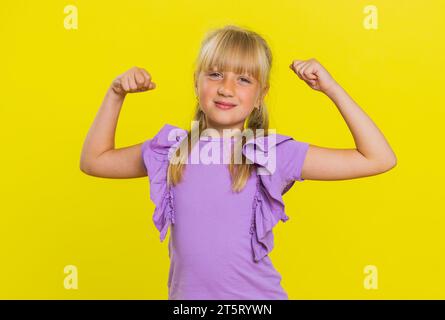 Je suis forte et en bonne santé. Jeune fille mignonne montrant des biceps et regardant confiant sentir la force de puissance pour se battre pour les droits, l'énergie pour gagner le succès gagner. Enfant blond préadolescent enfant sur fond jaune Banque D'Images