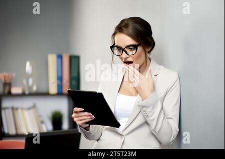 Jolie femme en tenue formelle et lunettes exprimant l'émotion surprise tout en utilisant la tablette. Lire des nouvelles choquantes ou des e-mails Réception d'un message important Banque D'Images