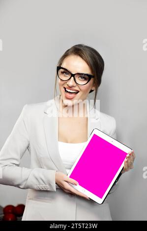 Belle jeune femme portant une veste et des lunettes à monture noire a excité l'expression faciale tout en démontrant quelque chose sur tablette numérique. Broche vide Banque D'Images