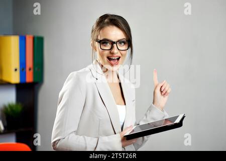 Jeune femme excitée pointant vers l'espace de copie libre sur le mur gris tout en tenant une tablette, portant des lunettes et des vêtements formels. Maquette pour publicité onlin Banque D'Images