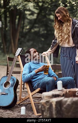 Un homme noir est assis sur une chaise de camping et tient un verre de bière, tandis que la femme blanche se tient à côté de lui et ils se sourient. Banque D'Images