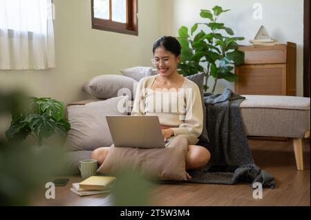 Une belle et heureuse femme asiatique discute avec ses amis sur Internet via son ordinateur portable tout en étant assise sur le sol à côté de son lit dans la chambre Banque D'Images