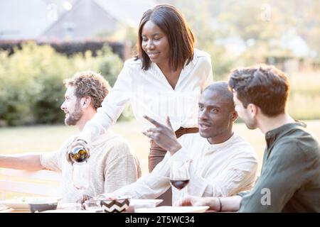 Au milieu des sons ambiants des rires et des verres clinquants, un groupe diversifié d'amis millénaires se réunit dans un jardin, célébrant leur lien autour d'un somptueux dîner. Le cœur du rassemblement est l'hôtesse gracieuse - la dame noire de la maison. Avec une bouteille de vin à la main, elle remplit le verre d'un invité, son geste incarnant l'esprit de générosité et de convivialité qui définit la soirée. Cheers to Friendship : l'hôtesse verse du vin au dîner multiracial Garden. Photo de haute qualité Banque D'Images