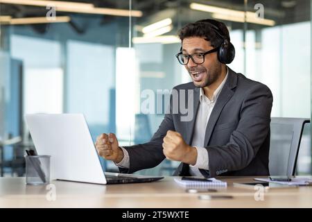 Un homme sur le lieu de travail à l'intérieur du bureau regarde un match de sport, un homme d'affaires dans des écouteurs et avec un ordinateur portable encourage sur le lieu de travail, un patron joyeux, célébrant une victoire, souriant. Banque D'Images