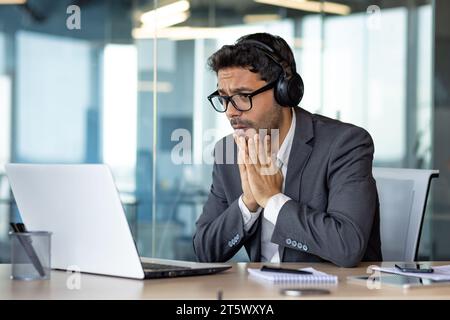 Homme regardant le match de sport à l'intérieur du bureau au travail, acclamations, homme d'affaires dans les écouteurs et ordinateur portable assis à table, malheureux avec le résultat triste et déçu. Banque D'Images