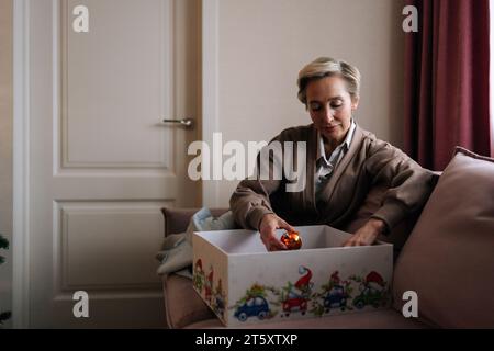 Portrait d'une femme élégante d'âge moyen choisissant des boules festives d'arbre de Noël dans une boîte assise sur un canapé. Préparation pour les vacances, décore l'arbre de Noël pour Banque D'Images