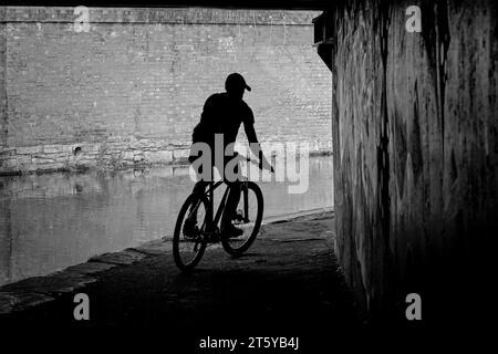 Un cycliste masculin se défile contre un mur de briques alors qu'il roule sous un pont le long d'un canal étroit à Huddersfield, Yorkshire, Angleterre, Royaume-Uni. Banque D'Images