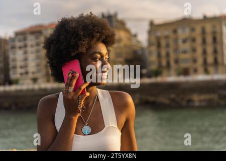 Portrait rapproché d'une jeune femme africaine distraite et souriante utilisant le téléphone près de la mer au coucher du soleil Banque D'Images