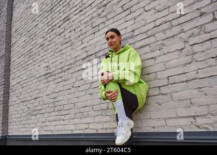 élégant jeune sportswoman à capuche couleur citron vert et leggings s'étendant près du mur gris à l'extérieur Banque D'Images