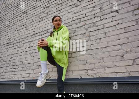 élégant jeune sportswoman à capuche couleur citron et leggings s'exerçant près du mur gris à l'extérieur Banque D'Images