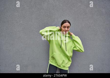 brunette femme dans le sweat à capuche couleur citron tendance ajustant la capuche et debout près du mur de béton gris Banque D'Images