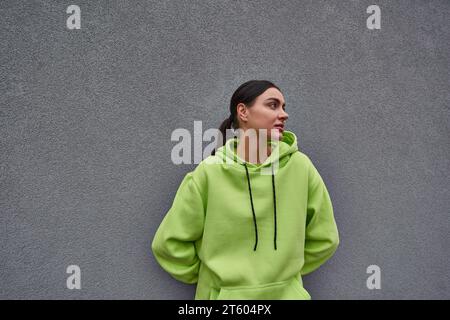 brunette jeune femme en sweat à capuche tendance couleur citron debout près du mur de béton gris à l'extérieur Banque D'Images