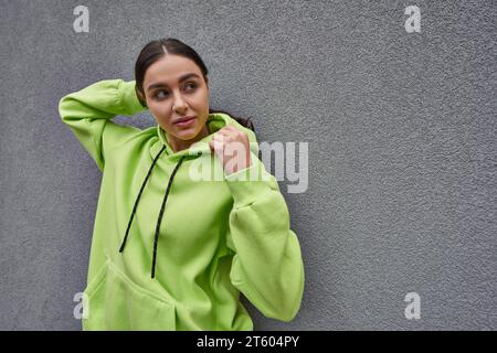 brunette jeune femme en sweat à capuche couleur citron à la mode posant tout en se tenant près du mur de béton gris Banque D'Images