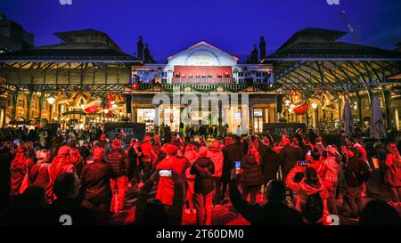 Londres, Royaume-Uni. 07 novembre 2023. Les gens, baignés de lumière rouge, regardent le London International Gospel Choir se produire sur le balcon du pub Punch and Judy. Nicole Scherzinger, Jamie Laing, Sophie Habboo, la troupe de SIX The musical et le London International Gospel Choir allument les lumières de Noël de Covent Garden sur la Piazza. Les lumières de Noël de Covent Garden présentent un nouveau schéma décoratif - le premier en près d'une décennie - avec 40 cloches gigantesques avec des arcs, 12 boules géantes et 8 boules de miroir tournantes. Crédit : Imageplotter/Alamy Live News Banque D'Images