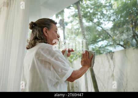 Femme âgée nettoyant les fenêtres dans sa maison avec du détergent en aérosol Banque D'Images